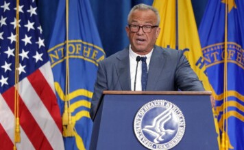 Older man in a suit speaks at a podium with the U.S. flag and department banners behind him.