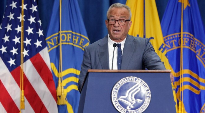 Older man in a suit speaks at a podium with the U.S. flag and department banners behind him.