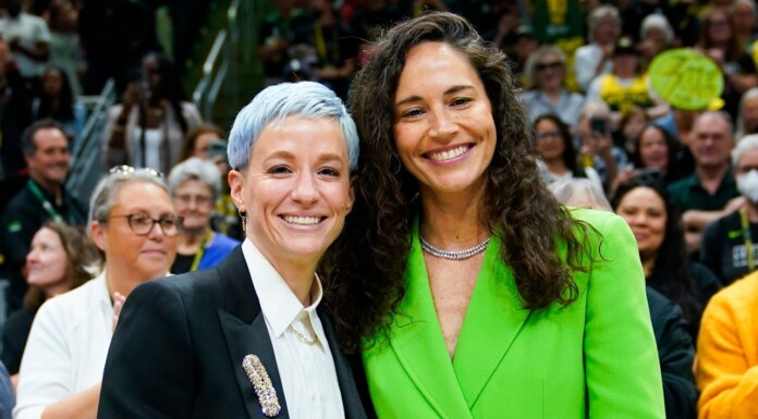 Two women posing together at a crowded event; one with short blue hair in a black blazer, the other in a bright green blazer.