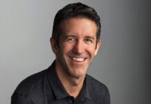 Professional head-and-shoulders portrait of a smiling man in a dark polo against a gray gradient background.