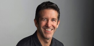 Professional head-and-shoulders portrait of a smiling man in a dark polo against a gray gradient background.