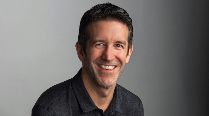Professional head-and-shoulders portrait of a smiling man in a dark polo against a gray gradient background.