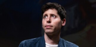 Young man with short dark hair wearing a blue jacket and white shirt looks upward against a dark background.