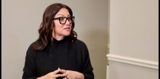 Woman with dark hair and black glasses in a black sweater, speaking with hands clasped in front of her.