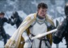 Knight in white armor with a gold-embroidered cape faces an opponent in a snowy battlefield, sword drawn.