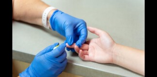 Healthcare worker in blue gloves pricking a patient's finger for a blood test or sample collection at a clinical desk area