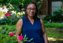 Portrait of a Black woman with glasses standing in a garden, wearing a dark blue sleeveless blouse, with pink roses in the foreground.