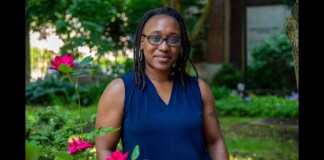 Portrait of a Black woman with glasses standing in a garden, wearing a dark blue sleeveless blouse, with pink roses in the foreground.