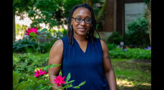 Portrait of a Black woman with glasses standing in a garden, wearing a dark blue sleeveless blouse, with pink roses in the foreground.