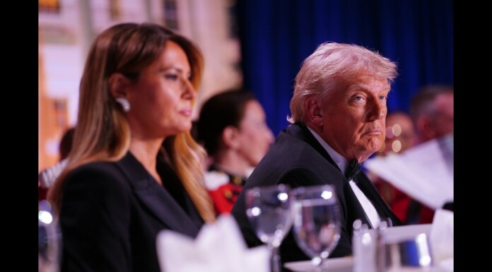 Trump Pulled From Correspondents’ Dinner Amid Reports of Gunfire, Suspect in Custody Side-profile of an older man with white hair in a dark tuxedo at a formal dinner, seated at a table with wine glasses, next to a woman, with a blurred background.