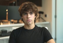 Young man with tousled brown hair wearing a black t‑shirt sits in an office, looking at the camera.