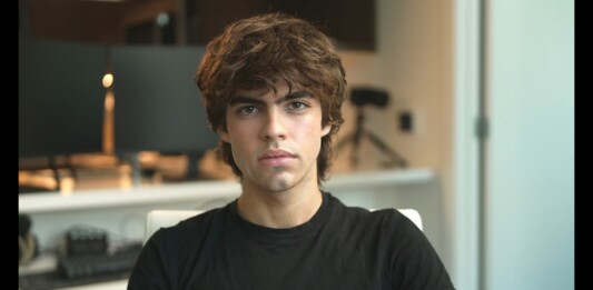 Young man with tousled brown hair wearing a black t‑shirt sits in an office, looking at the camera.