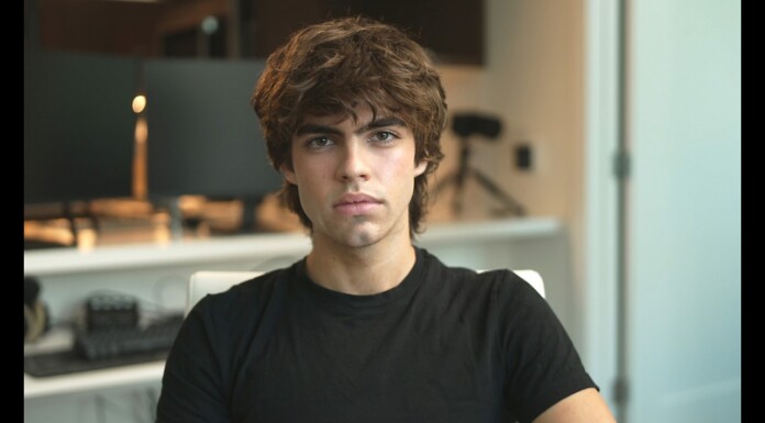 Viral “Gay Is a Compliment” Claim Ignites Debate on Stereotypes in Looksmaxxing Culture Young man with tousled brown hair wearing a black t‑shirt sits in an office, looking at the camera.