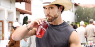 Young man in a straw hat sipping a red beverage through a green straw while holding a sandwich at an outdoor market/event.