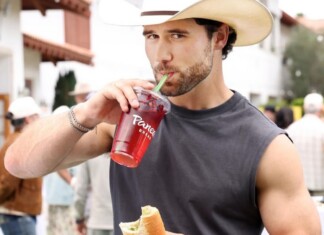 Young man in a straw hat sipping a red beverage through a green straw while holding a sandwich at an outdoor market/event.