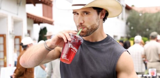 Young man in a straw hat sipping a red beverage through a green straw while holding a sandwich at an outdoor market/event.