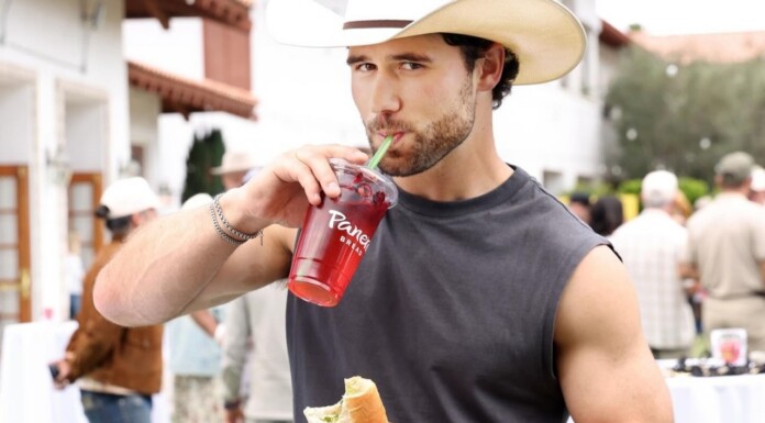 Young man in a straw hat sipping a red beverage through a green straw while holding a sandwich at an outdoor market/event.