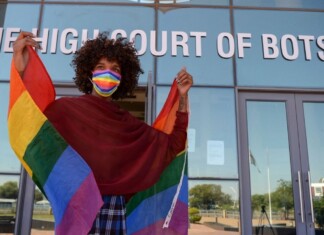 Botswana Repeals Anti-Sodomy Law, Marking Major LGBTQ+ Legal Milestone Person with curly hair wears a rainbow mask and cape-like Pride flag outside The High Court of Botswana, smiling at the camera.