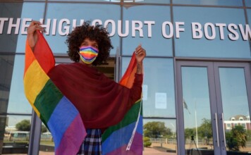 Person with curly hair wears a rainbow mask and cape-like Pride flag outside The High Court of Botswana, smiling at the camera.