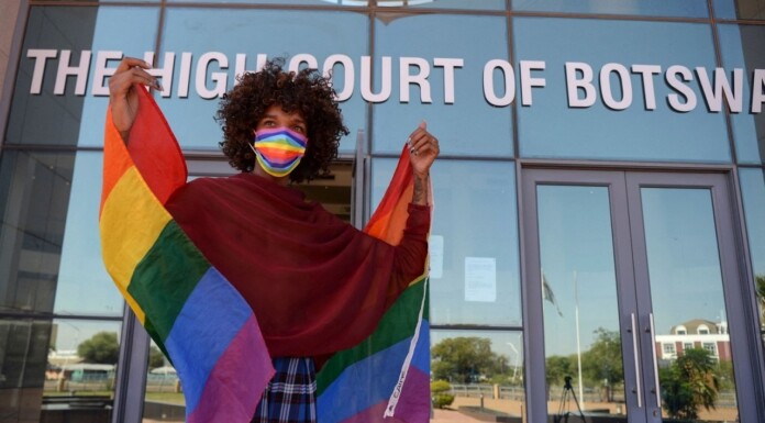Person with curly hair wears a rainbow mask and cape-like Pride flag outside The High Court of Botswana, smiling at the camera.