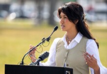 Woman in a beige vest and white shirt speaks at a podium with multiple microphones outdoors. (Informative: a speaker giving a speech)