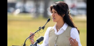 Woman in a beige vest and white shirt speaks at a podium with multiple microphones outdoors. (Informative: a speaker giving a speech)