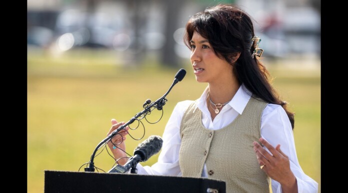 Woman in a beige vest and white shirt speaks at a podium with multiple microphones outdoors. (Informative: a speaker giving a speech)