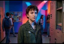 Young student with short curly hair wearing a green jacket stands in a colorful school hallway with lockers on both sides.