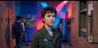 Young student with short curly hair wearing a green jacket stands in a colorful school hallway with lockers on both sides.