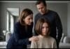 Mother and father lean in to comfort their somber daughter seated in a modern living room, hand on her shoulder.",