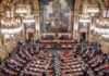A grand legislative chamber with ornate gold chandeliers, marble walls, and rows of desks as lawmakers gather for session.