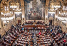 A grand legislative chamber with ornate gold chandeliers, marble walls, and rows of desks as lawmakers gather for session.