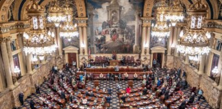 A grand legislative chamber with ornate gold chandeliers, marble walls, and rows of desks as lawmakers gather for session.