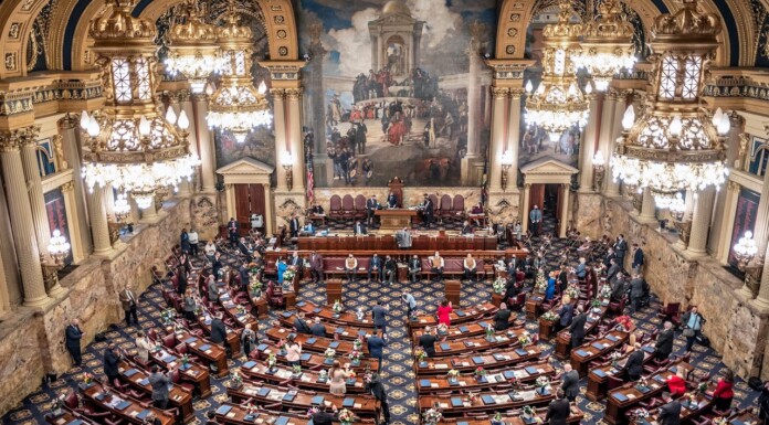A grand legislative chamber with ornate gold chandeliers, marble walls, and rows of desks as lawmakers gather for session.