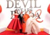 Group of four on a red carpet posing in glamorous outfits, with a large 'Devil Wears Prada' backdrop and oversized red heels on either side.