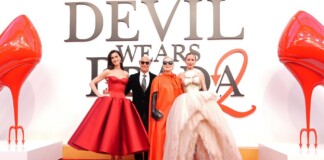 Group of four on a red carpet posing in glamorous outfits, with a large 'Devil Wears Prada' backdrop and oversized red heels on either side.
