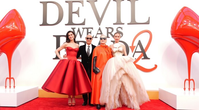 Group of four on a red carpet posing in glamorous outfits, with a large 'Devil Wears Prada' backdrop and oversized red heels on either side.