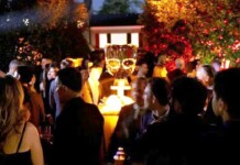 Crowd of people at a nighttime outdoor party around a glowing champagne tower centerpiece on a pedestal outside.