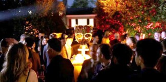 Crowd of people at a nighttime outdoor party around a glowing champagne tower centerpiece on a pedestal outside.