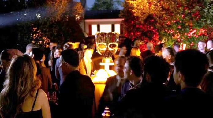Crowd of people at a nighttime outdoor party around a glowing champagne tower centerpiece on a pedestal outside.