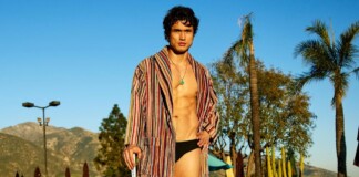 Man in a striped robe over black swim briefs posing poolside at a sunny resort with palm trees in the background