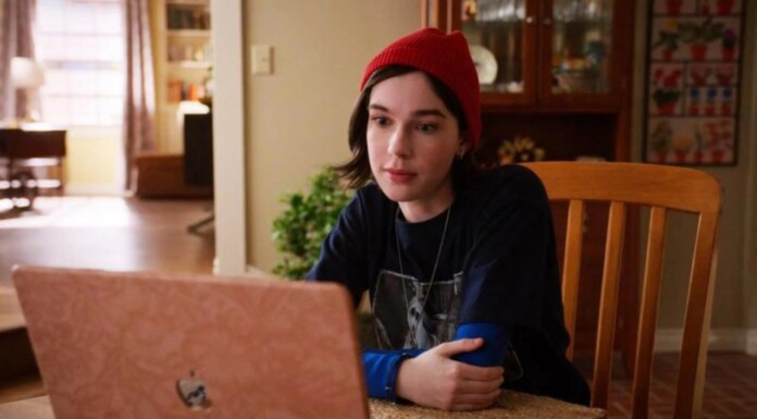 Malcolm in the Middle Reboot Has A Nonbinary Character Kelly Young person wearing a red beanie sits at a dining table indoors, using a pink laptop and looking at the screen.a0