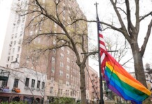 Rainbow Pride flag and American flag flutter on a flagpole in a city street, with bare trees and tall buildings in the background.