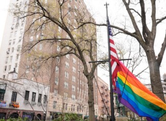 Rainbow Pride flag and American flag flutter on a flagpole in a city street, with bare trees and tall buildings in the background.