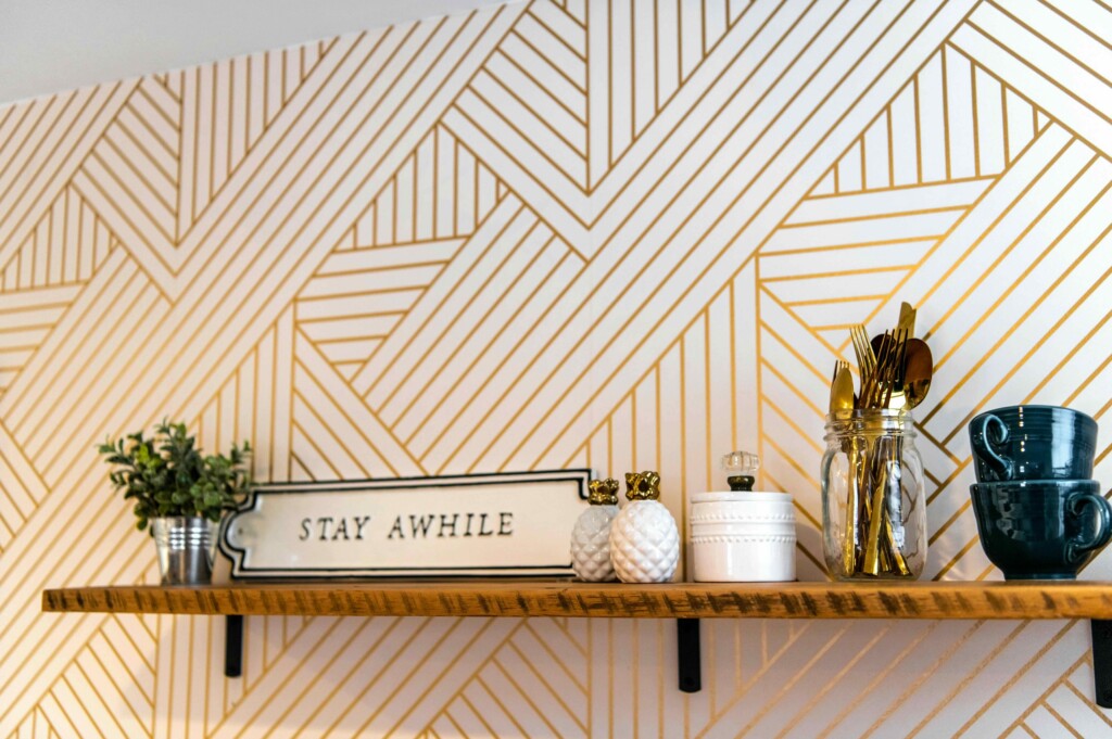 Shelf display with glass jars and gold utensils in front of a gold chevron wall pattern, plus a small plant and a 'STAY AWHILE' sign nearby.