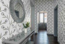 Decorative entry hall with large circular textured mirror on left, patterned wallpaper, glass console table and vase of white flowers; doorway to a bright kitchen beyond.