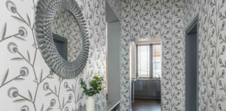 Decorative entry hall with large circular textured mirror on left, patterned wallpaper, glass console table and vase of white flowers; doorway to a bright kitchen beyond.