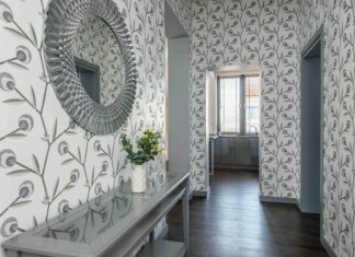 Decorative entry hall with large circular textured mirror on left, patterned wallpaper, glass console table and vase of white flowers; doorway to a bright kitchen beyond.