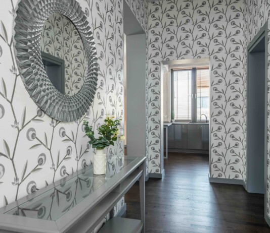 Decorative entry hall with large circular textured mirror on left, patterned wallpaper, glass console table and vase of white flowers; doorway to a bright kitchen beyond.