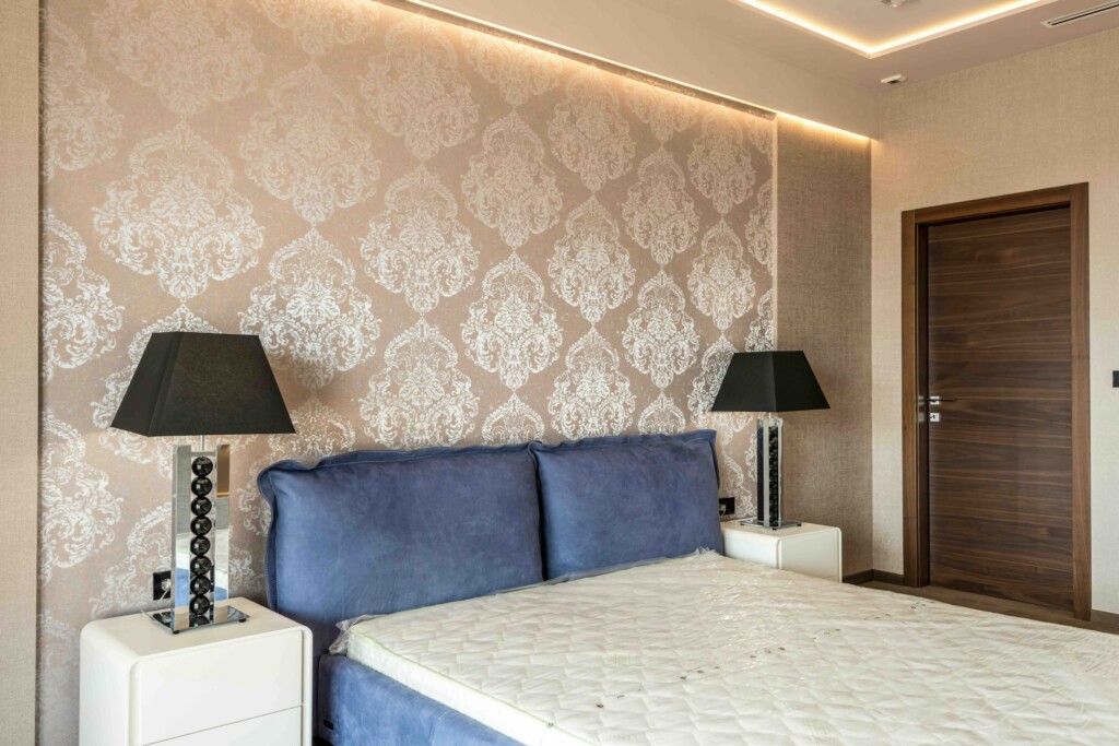 Hotel bedroom with damask patterned wallpaper, blue headboard pillows, black lamps, white nightstands, and a wooden door on the right